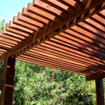 Close up of finished wooden Pergola in front of green bushes with pink flowers